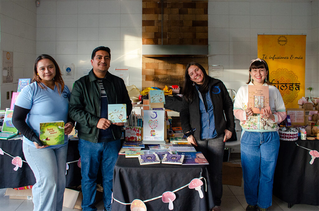 Cuatro personas (tres mujeres, un hombre) sonriendo detrás de una mesa llena de libros y decorada con una guirnalda de setas en un evento comunitario de Gota de Lluvia. La mujer de la izquierda sostiene un libro verde titulado "Dulce y Suave", el hombre sostiene el libro "nido", y la mujer de la derecha sostiene el libro "Dulce Pincelada de mi nostalgia". La mesa tiene otros carteles, incluido uno para té, infusiones e hamsa, y un cartel central de un libro ilustrado.