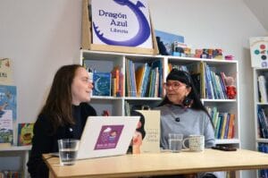 Cecilia Lagos, de Gota de Lluvia, entrevista a la escritora Malucha Pinto en la Librería Dragón Azul en Valdivia. Están sentadas a una mesa de madera con micrófonos, vasos con agua y un computador portátil que exhibe el sticker del logo de Gota de Lluvia. Malucha Pinto sostiene su libro Cartas para Tomás, de Editorial Catalonia, mientras conversan sonrientes. De fondo se aprecian estanterías repletas de libros y el letrero "Dragón Azul Librería".