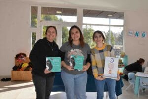 Fotografía de Loreto San Martín, Diana Rivas y Valentina Lobos sonriendo durante un evento de fomento lector para infancias en un espacio de juego luminoso. Dos de ellas sostienen el libro "Poeta de las cosas simples", mientras que la tercera sostiene un cartel informativo enmarcado. Al fondo, se observan niños y un cofre de juguetes.